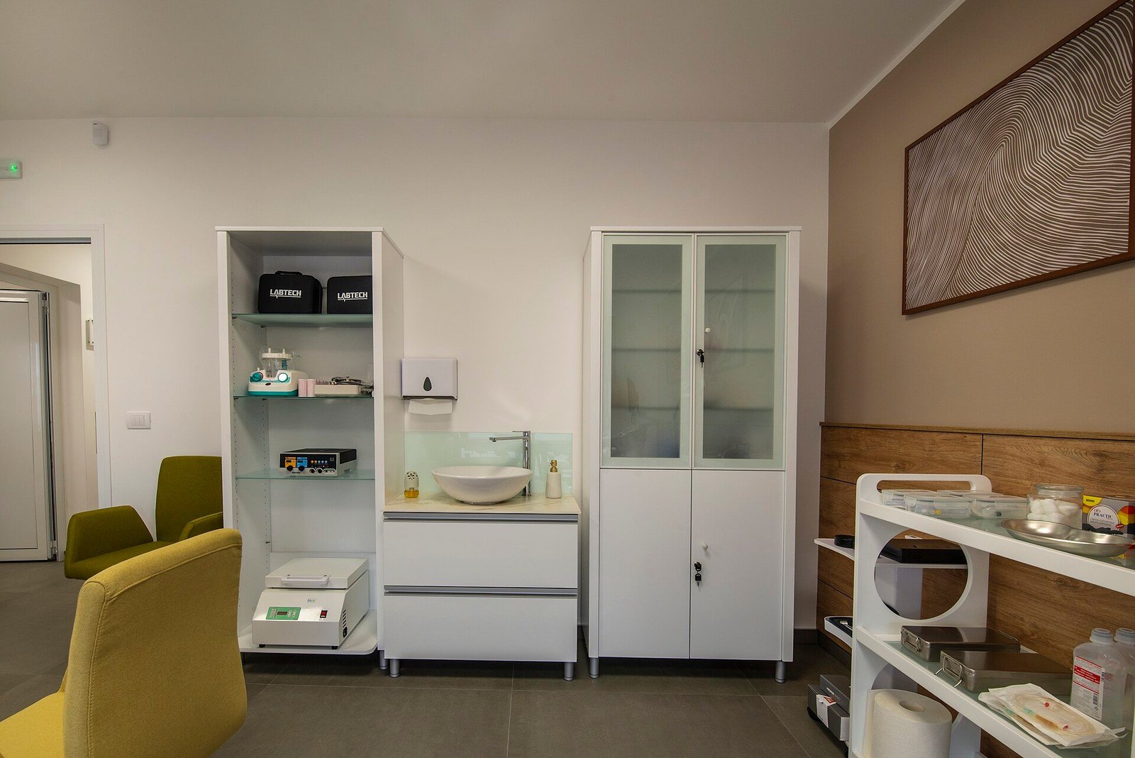Interior of a modern medical aesthetic clinic room featuring white storage cabinets with glass shelves holding medical equipment, a wall-mounted hand-wash basin, a locked supply cabinet, a white treatment trolley with skincare products, and olive-green waiting chairs against neutral-toned walls with